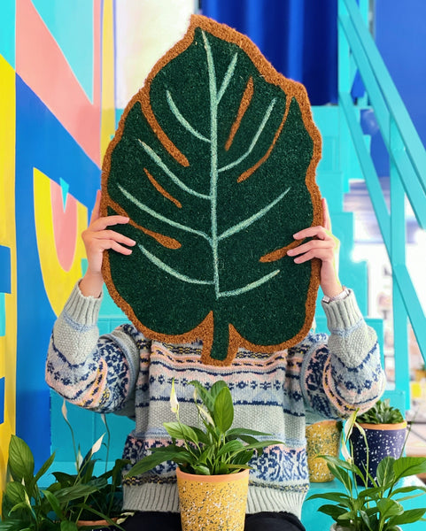 Chica sujetando un felpudo con forma de hoja de monstera rodeada de macetas y plantas