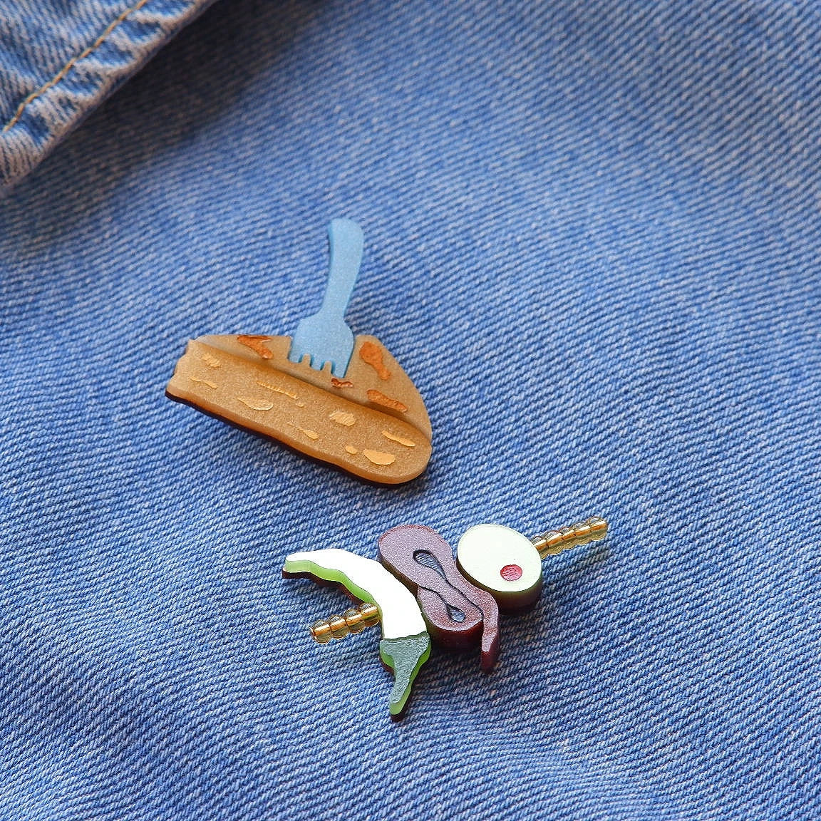 pin de un pincho de tortilla en metacrilato con un tenedor pinchandolo y pin de una gilda con cuentas doradas de cristal de la artista mitumi, colocados en una chaqueta vaquera