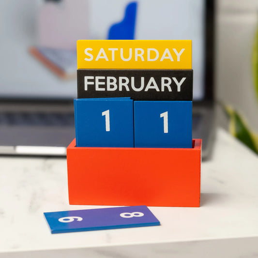 calendario pequeño de mesa multicolor con una caja en color rojo, piezas con números en color azul, el mes de febrero en color negro y el día de la semana Sábado en color amarillo