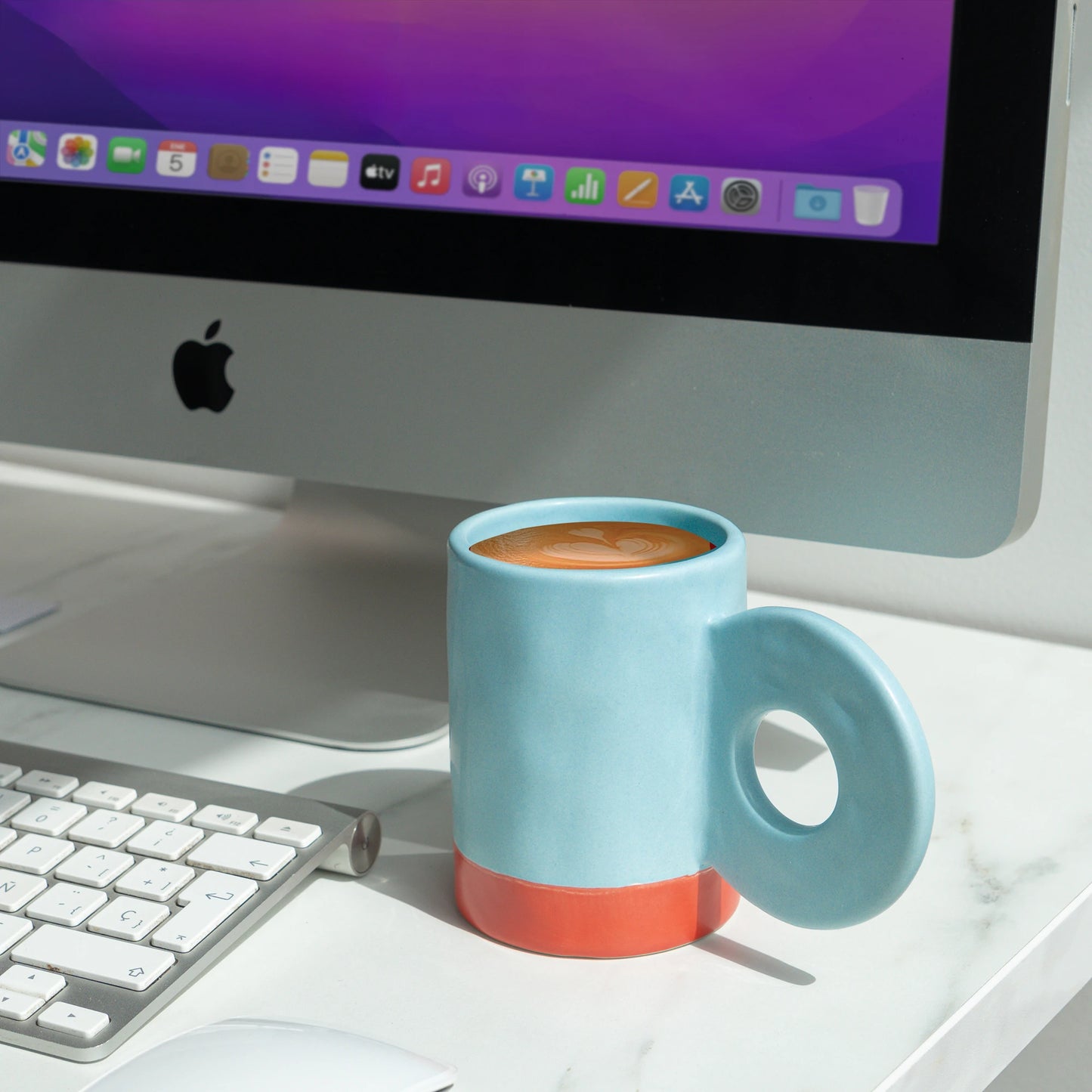 taza de cerámica de color celeste de Helio Ferretti llena de café con la base de color rojo, con asa grande en forma de círculo  colocada en una mesa en frente de un ordenador