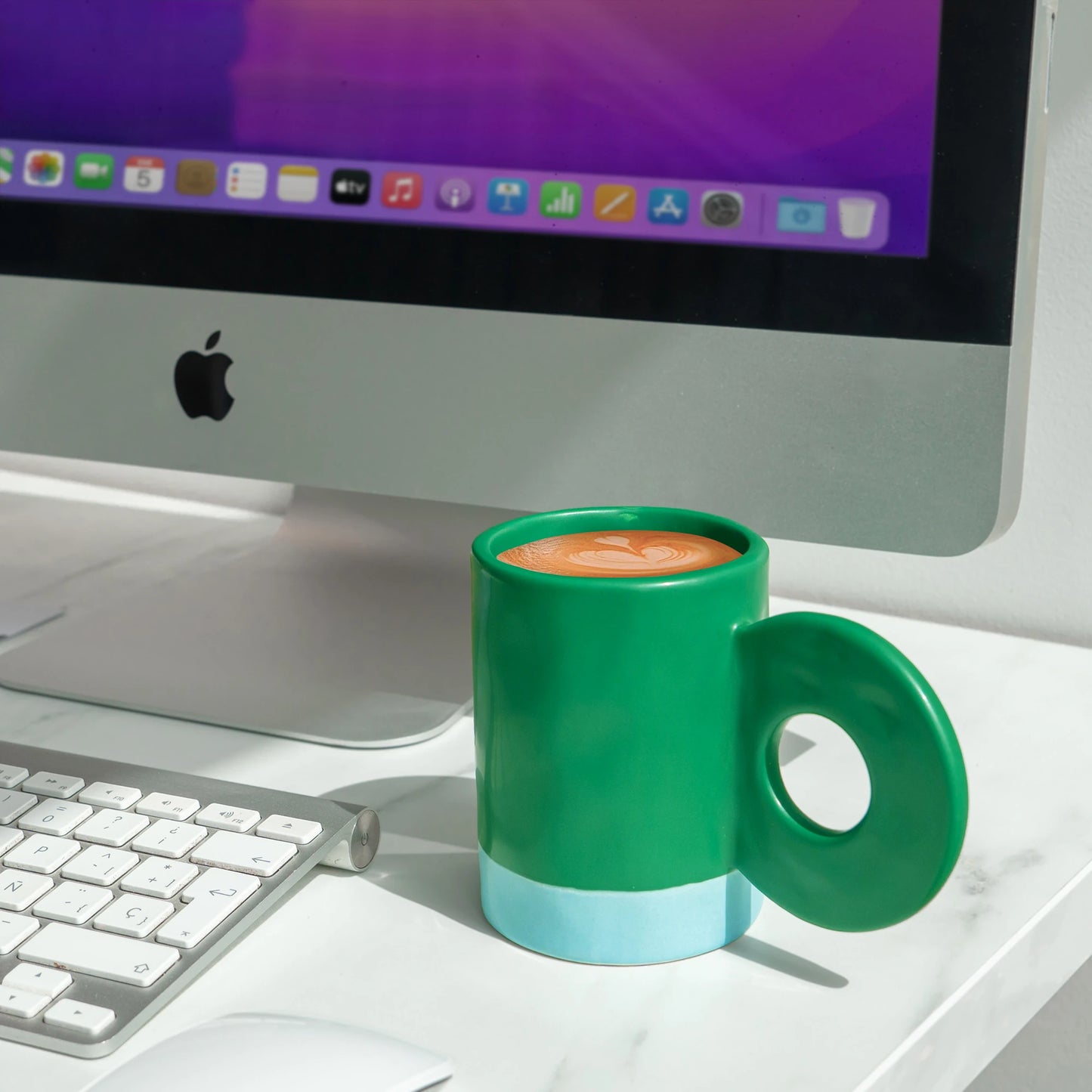 taza de cerámica de color verde de Helio Ferretti llena de café con la base de color azul celeste, con asa grande en forma de círculo  colocada en una mesa en frente de un ordenador