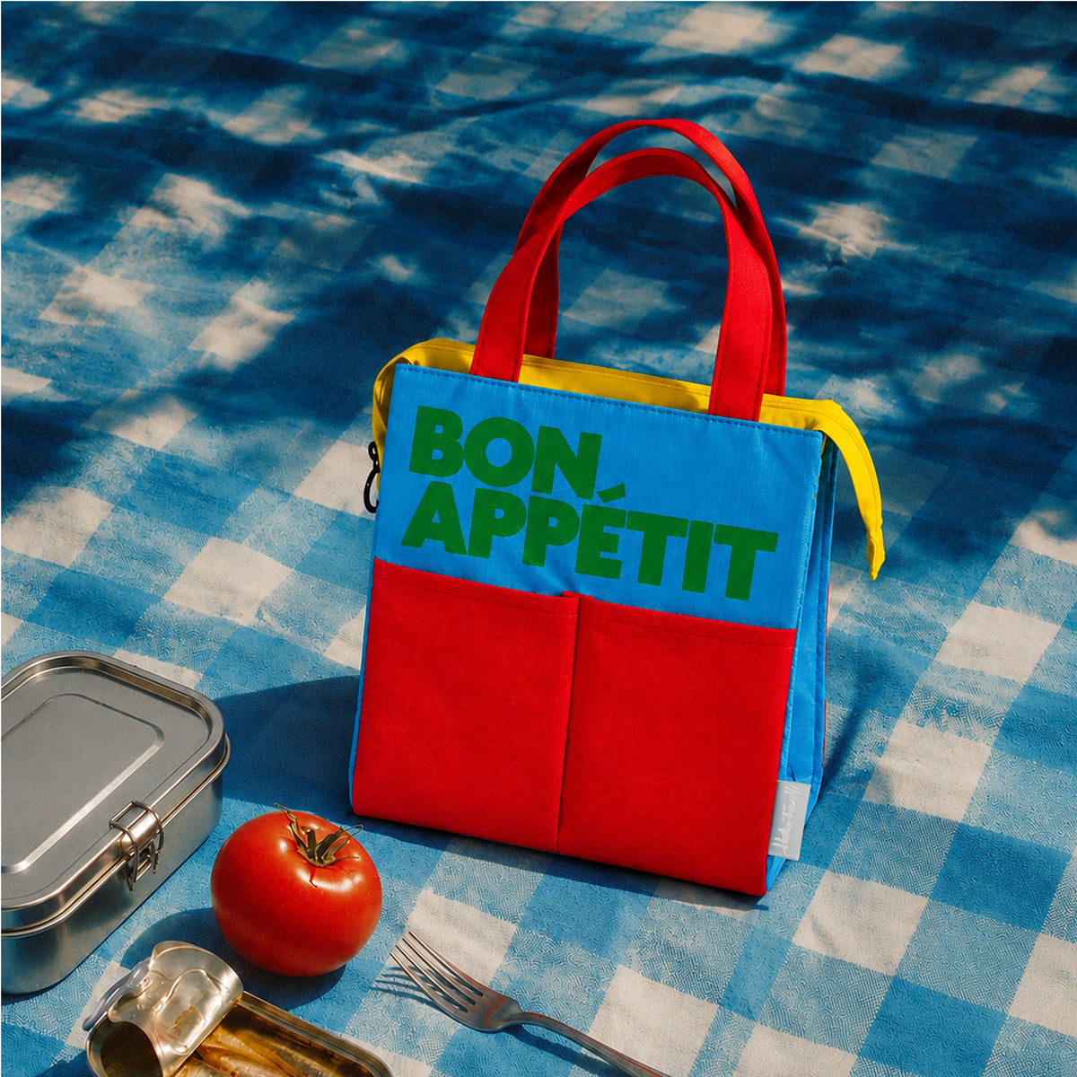 Bolsa isotérmica azul Bon Appétit de Helio Ferretti con asas y bolsillos rojos, sobre un mantel de picnic junto a una fiambrera.

