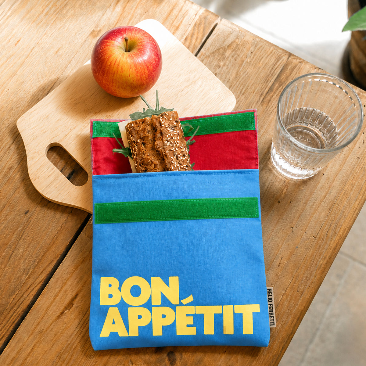 Bolsa de almuerzo azul Bon Appétit de Helio Ferretti, abierta con un bocadillo en su interior sobre una mesa de madera.