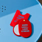 Espejo de bolsillo rojo More Chill, Less Stress de Helio Ferretti, abierto junto a su funda sobre una silla azul.

