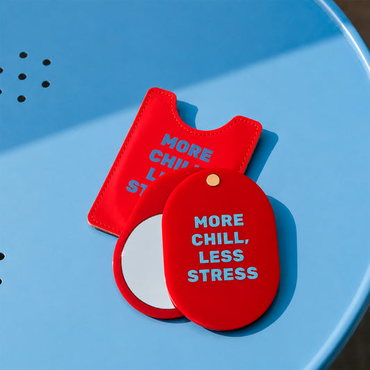 Espejo de bolsillo rojo More Chill, Less Stress de Helio Ferretti, abierto junto a su funda sobre una silla azul.
