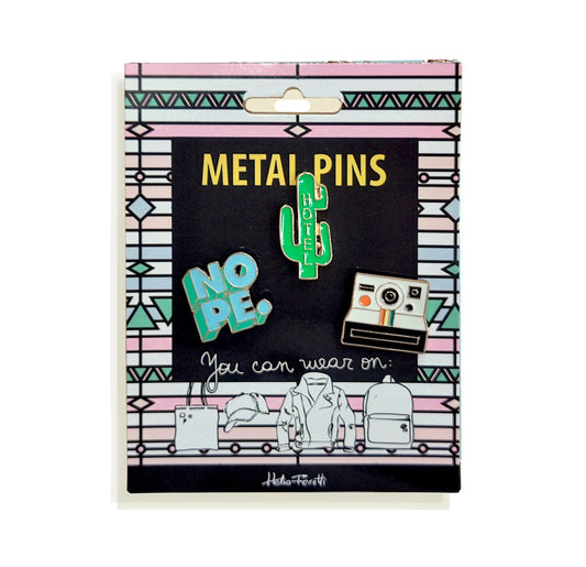 conjunto de tres pines de metal de Helio Ferretti en una tarjeta de cartón de colores rosa, azul y blanco con un patrón de figuras geométricas, con un diseño de una cámara polaroid, un cactus con la palabra HOTEL y la palabra NOPE.