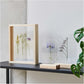 soporte de madera con panel acrílico transparente con la base en color madera colocado sobre una mesa y con flores dentro haciendo un efecto flotante