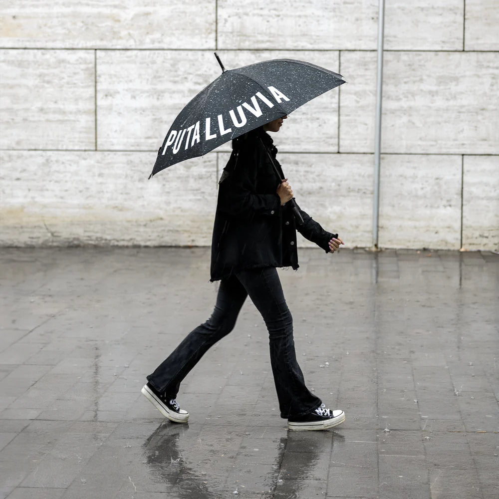 persona caminando por la calle lluviosa de lado con el paraguas de Fisura negro con el texto PUTA LLUVIA escrito en blanco