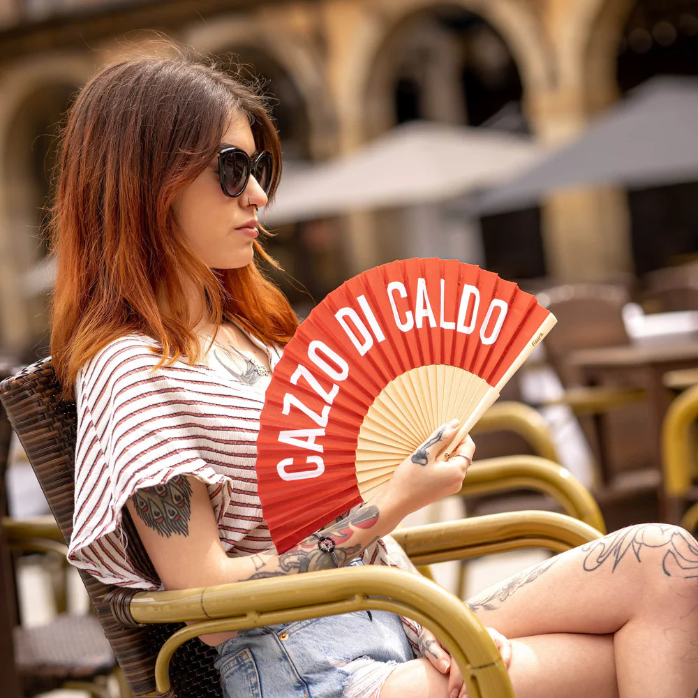 Chica sentada en una silla aguantando el abanico con el texto "Cazzo di caldo".