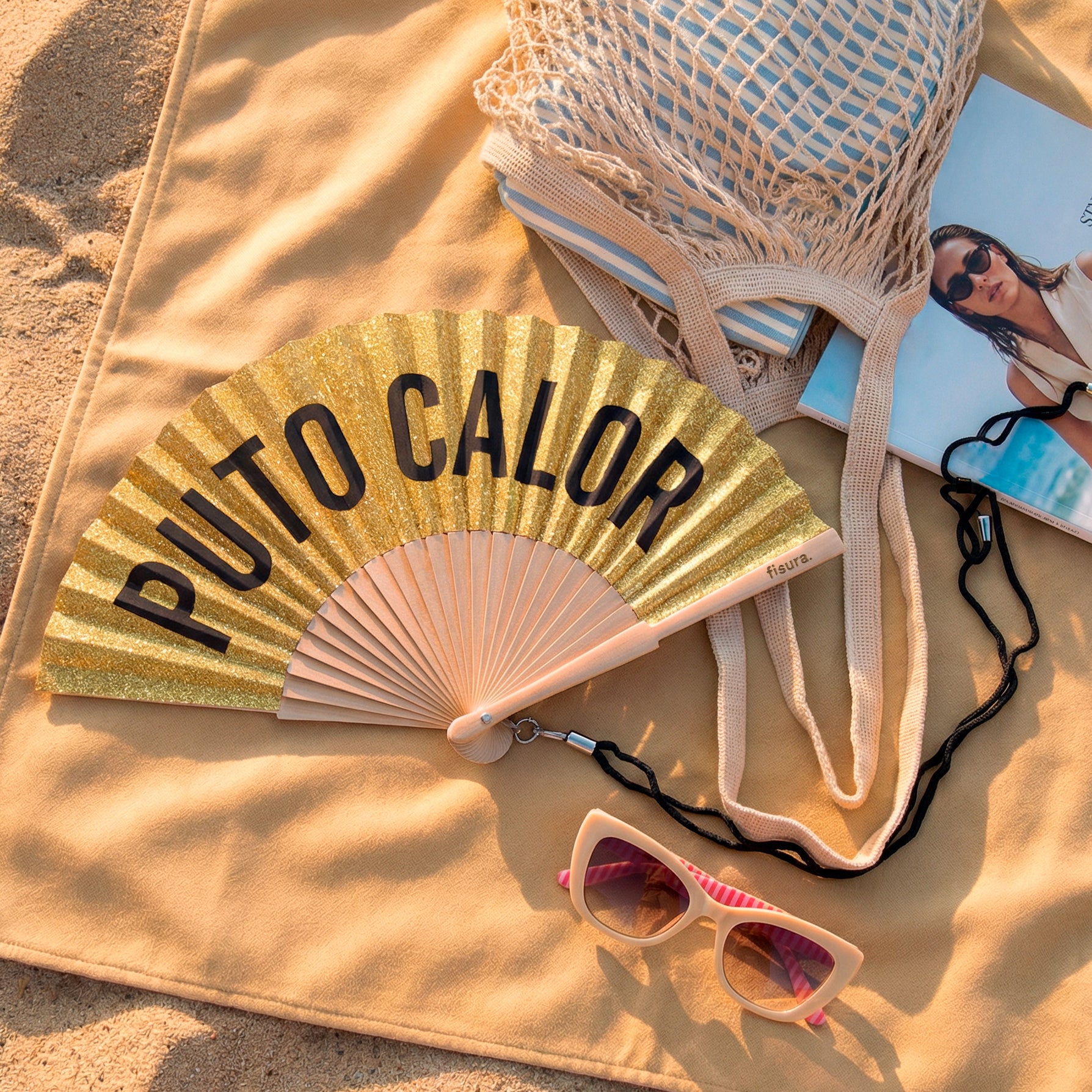 abanico de la marca fisura con purpurina dorada y el texto "PUTO CALOR" en color negro, sobre una toalla en la playa al lado de una bolsa y unas gafas de sol
