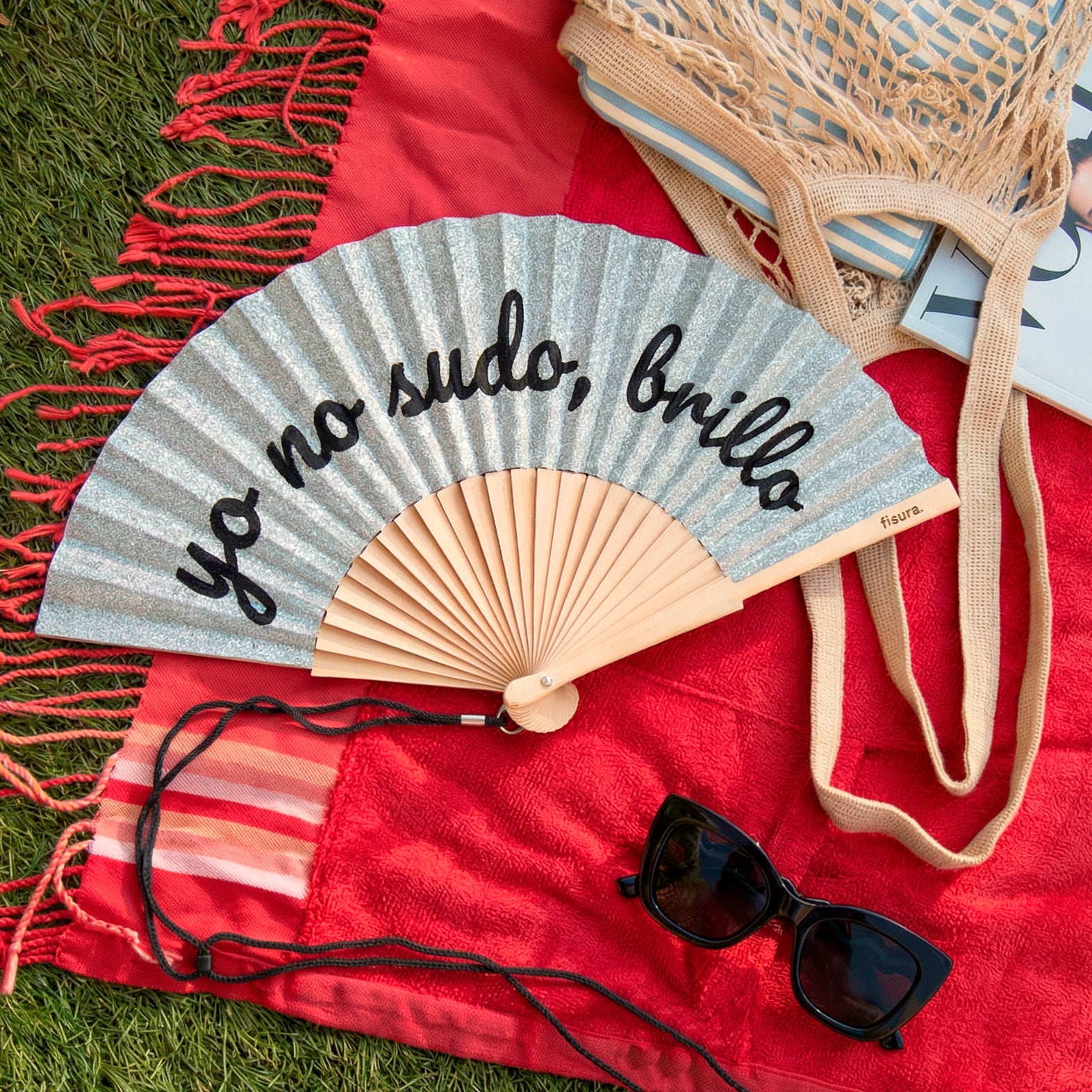 abanico de la marca fisura con purpurina plateada y el texto "yo no sudo, brillo" en color negro sobre una toalla en color rojo con una bolsa y unas gafas de sol