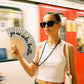 chica sujetando el abanico de la marca fisura con purpurina plateada y el texto "yo no sudo, brillo" en color negro con la cuerda negra colgada al cuello y en un metro 
