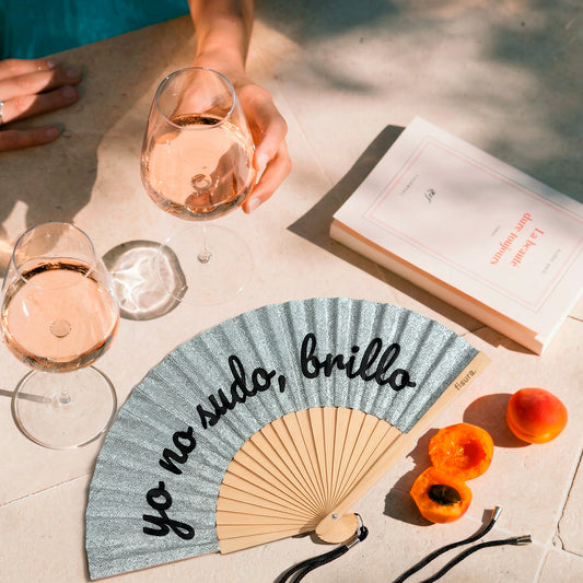 abanico de la marca fisura con purpurina plateada y el texto "yo no sudo, brillo" en color negro, sobre una mesa con libros, bebidas y fruta