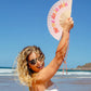chica posando con el abanico de la marca fisura en color melocotón y con la frase "No me da la vida" con las letras en colores rojo, naranja, amarillo y rosa, en la playa y con la cuerda para colgarlo a la muñeca