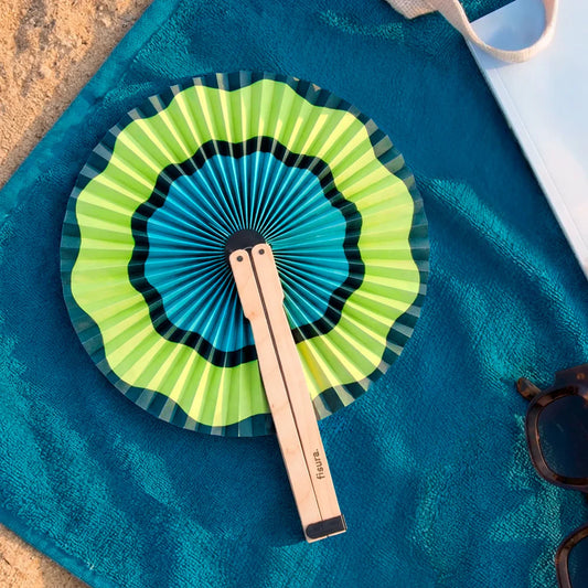Paipai plegable redondo de papel con estampado de ondas en tonos verdes y azules en una toalla en la playa