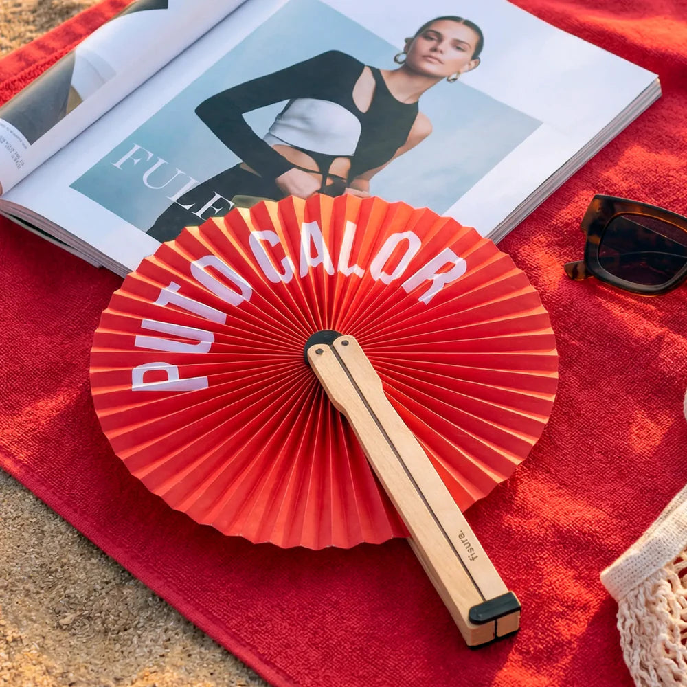 Paipai plegable redondo de papel en color rojo y con el texto PUTO CALOR en blanco sobre una toalla roja en la playa y con una revista y unas gafas de sol al lado