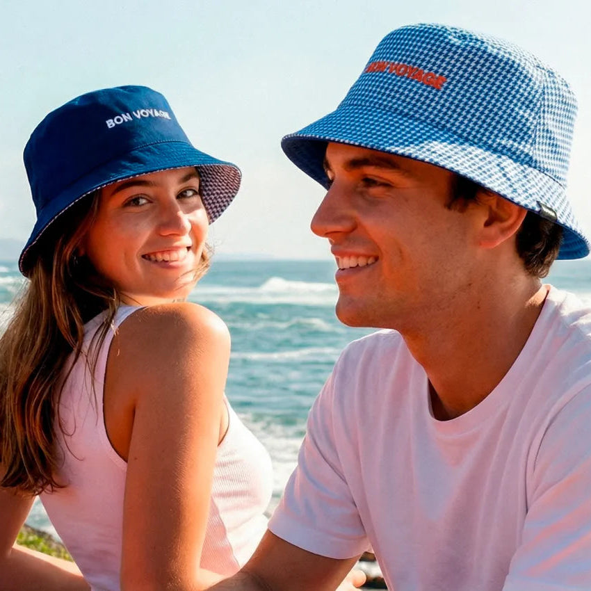 pareja con el chico llevando el gorro de Fisura reversible a cuadros azul marino y blanco y el texto BON VOYAGE en color naranja, y la chica con el gorro BON VOYAGE en color azul marino y las letras blancas