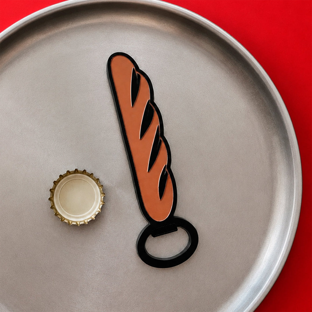 Abridor magnético con forma de barra de pan sobre un plato metálico en una mesa roja junto a una chapa