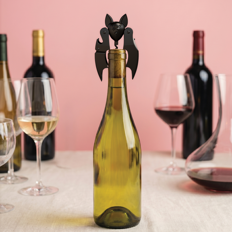 Sacacorchos Murciélago de Ototo colocado sobre una botella de vino blanco, con copas y otras botellas al fondo.