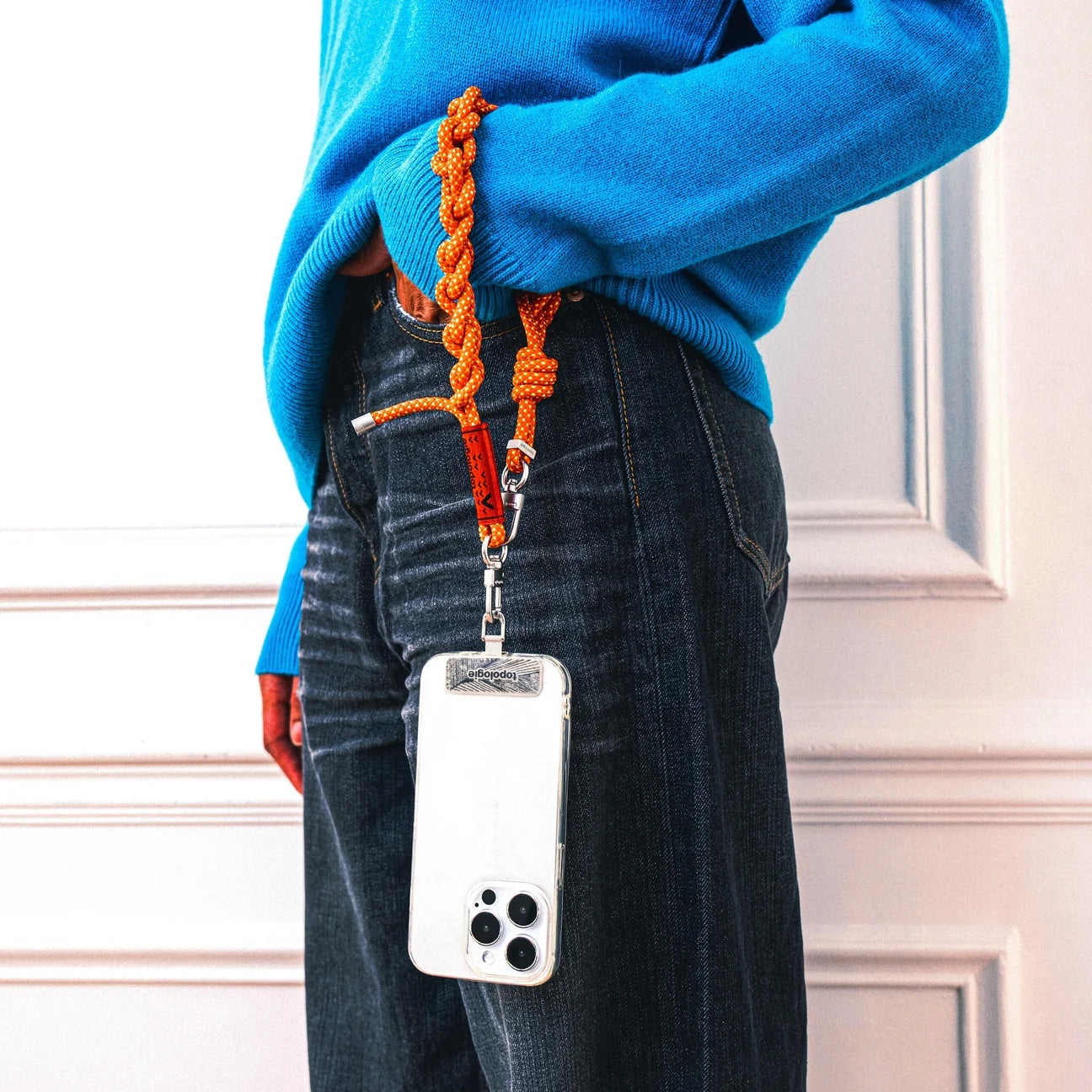 Chica con una correa de mano naranja enganchada al móvil con el adaptador para enganchar correas al móvil de Topologie con gancho de acero inoxidable