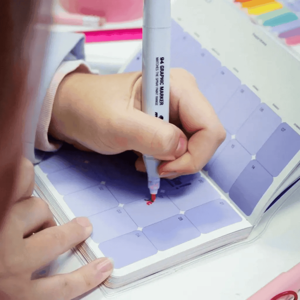 Mano escribiendo en agenda pocket Happimess con rotulador, mostrando la apertura flat-lay a 180°