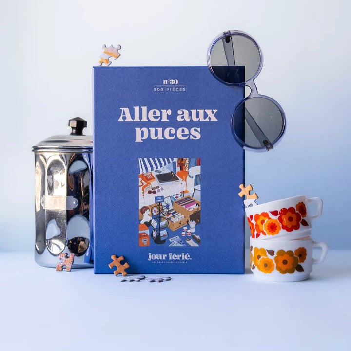Caja del puzzle de 500 piezas de color azul en la que pone "Aller aux puces" con decoracion de gafas, tazas y cafeteras de fondo. 