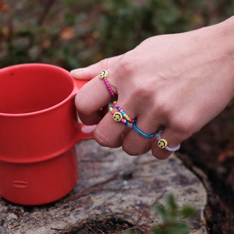 Foto de una mano con anillos de cuentas coloridas y con caritas sonrientes cogiendo una taza de café