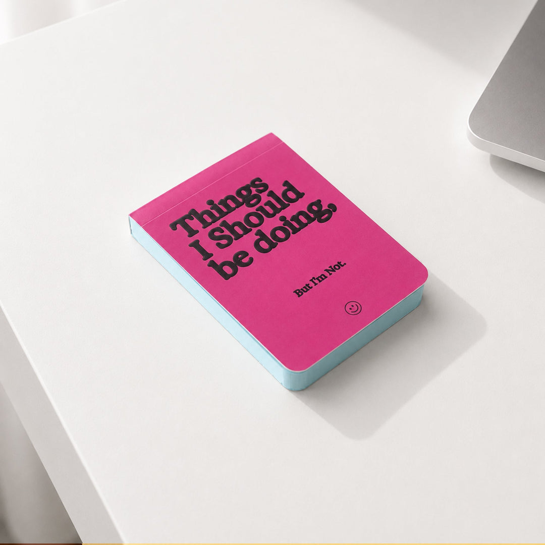 Mesa de trabajo con un bloc de notas con portada rosa fucsia y la frase Things I Should be doing, But I'm Not