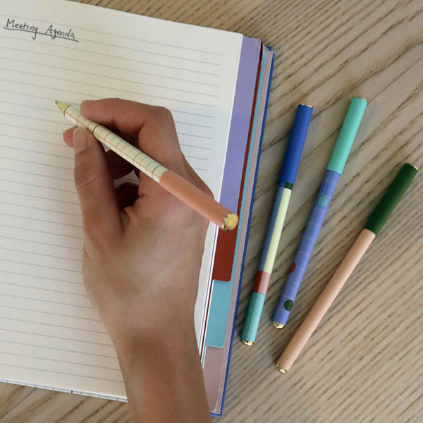 Mano escribiendo en una agenda a rayas con un boli metálico a cuadros y colores pastel, con otros tres bolígrafos a juego sobre la mesa