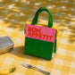 «Bolsa isotérmica Bon Appétit rosa y verde de Helio Ferretti sobre un mantel de cuadros amarillos, junto a una fiambrera y un tenedor.»