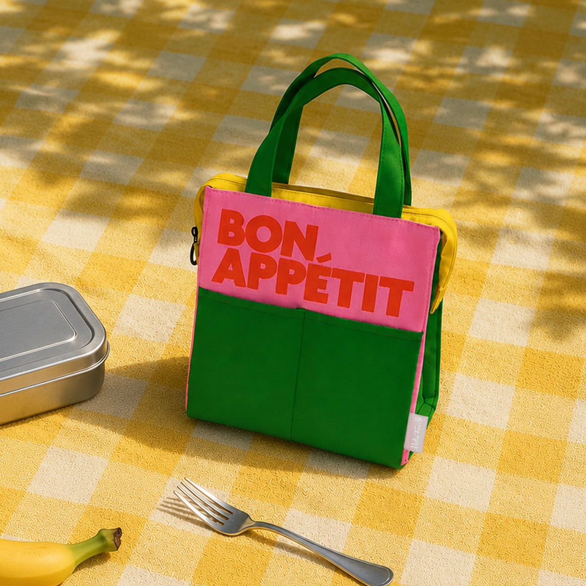 «Bolsa isotérmica Bon Appétit rosa y verde de Helio Ferretti sobre un mantel de cuadros amarillos, junto a una fiambrera y un tenedor.»