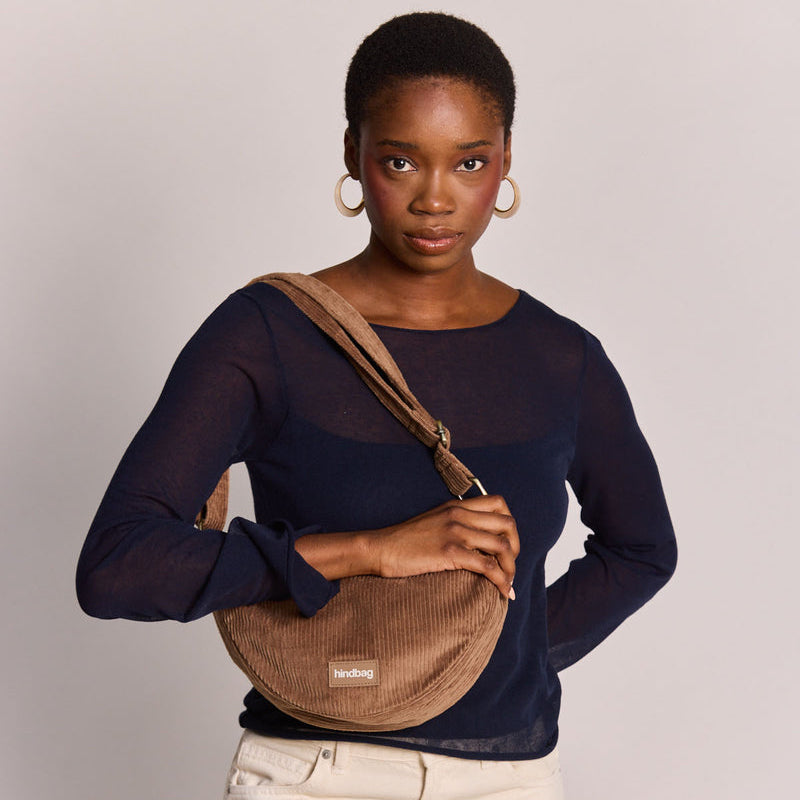Mujer de frente con pendientes de aro grandes y camiseta azul marino y el bolso de pana con forma de media luna en color marrón café con leche de la marca francesa Hindbag colgado al hombro