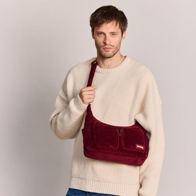 Hombre con jersey blanco y vaqueros con el bolso de pana color burdeos, dos bolsillos frontales con cremallera y correa ajustable de la marca francesa Hindbag 