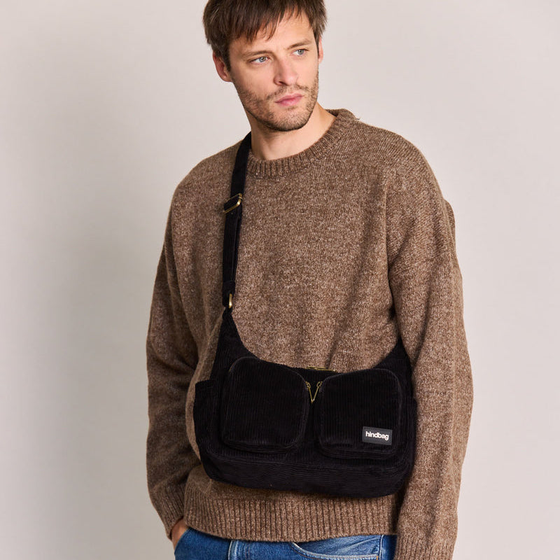 Hombre con jersey marrón y vaqueros y bolso de pana color negro con dos bolsillos frontales con cremallera y correa ajustable de la marca francesa Hindbag 