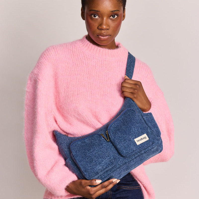 Mujer con jersey rosa chicle y vaqueros con bolso de tela vaquera azul con dos bolsillos frontales con cremallera de la marca francesa Hindbag colgado en bandolera