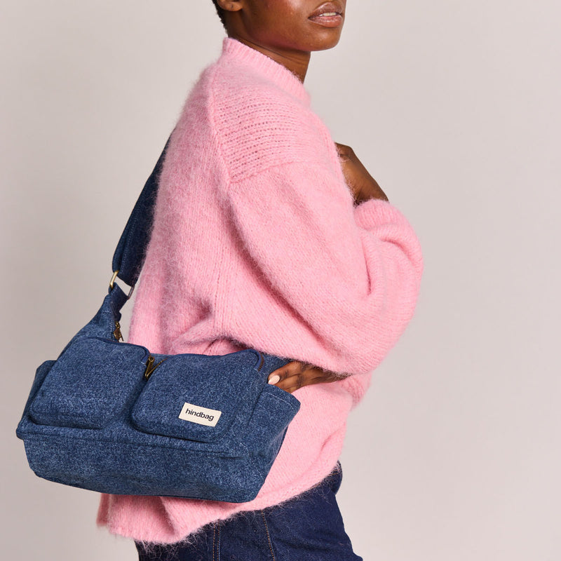 Mujer de perfil con vaqueros y jersey rosa con bolso de tela vaquera azul, dos bolsillos frontales con cremallera y correa ajustable de la marca francesa Hindbag colgado al hombro