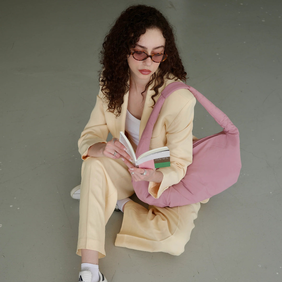 Mujer sentada en el suelo leyendo un libro con traje de chaqueta beige y bolso rosa acolchado de Lefrik