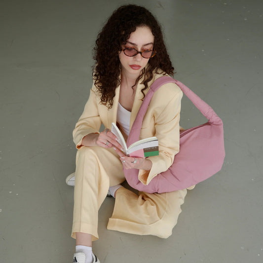 Mujer sentada en el suelo leyendo un libro con traje de chaqueta beige y bolso rosa acolchado de Lefrik