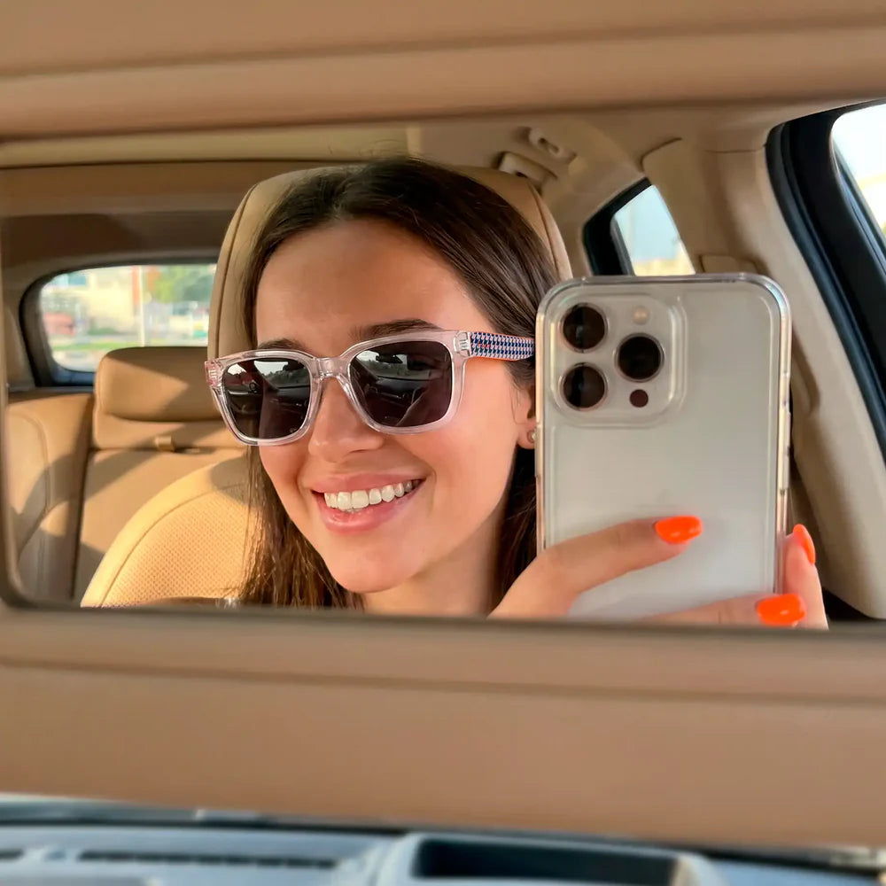 modelo posando con las gafas de sol de Fisura del modelo Bon Voyage cuadradas con la parte delantera transparente y los cristales en negro y las patillas con patrones geométricos en blanco y azul, haciéndose una foto en el espejo del coche