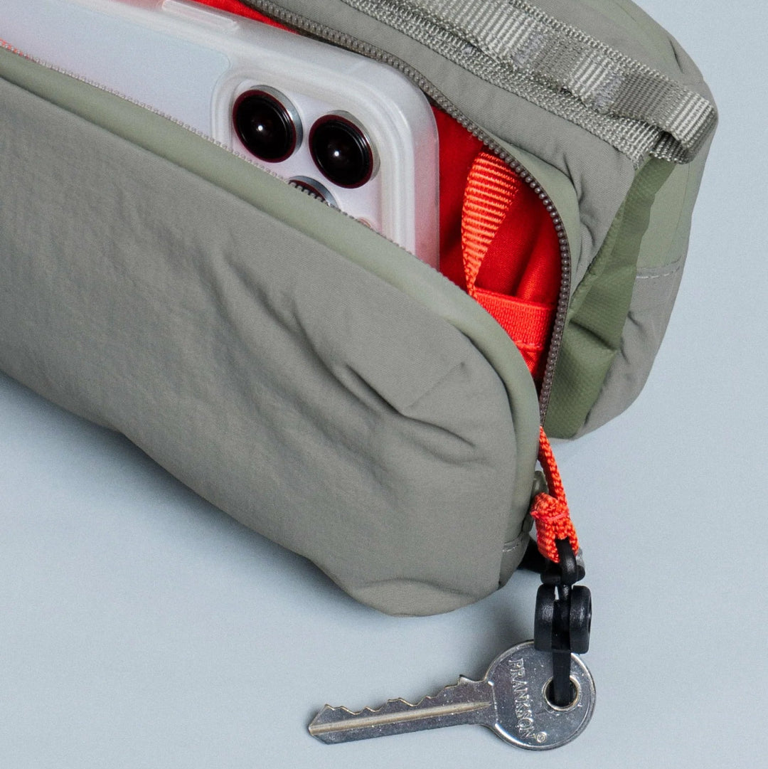 Detalle del bolso Bottle Sacoche Medium Topologie en color caqui con cremallera abierta, móvil en el interior y llave colgando del tirador
