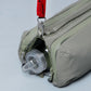 Detalle del compartimento inferior del bolso Bottle Sacoche Medium Topologie en color caqui con botella de agua asomando y correa Topologie roja enganchada
