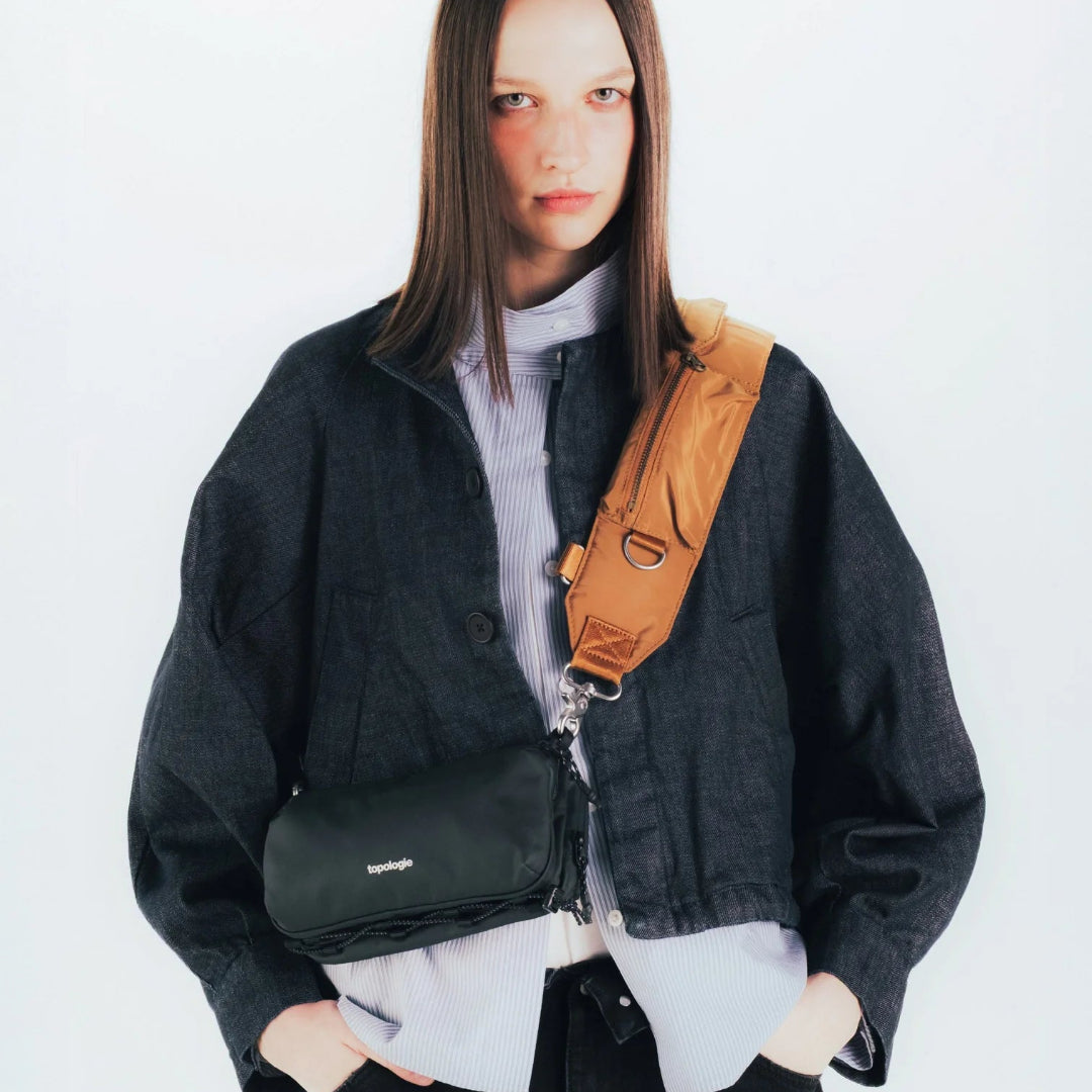 Modelo femenina con bolso Bottle Sacoche Medium Topologie negro cruzado al cuerpo con correa ancha en color camel, combinado con chaqueta vaquera oscura
