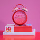 reloj retro de color rojo y con las manecillas rosas, con el texto better late than ugly sobre una pila de libros y un fondo rosa