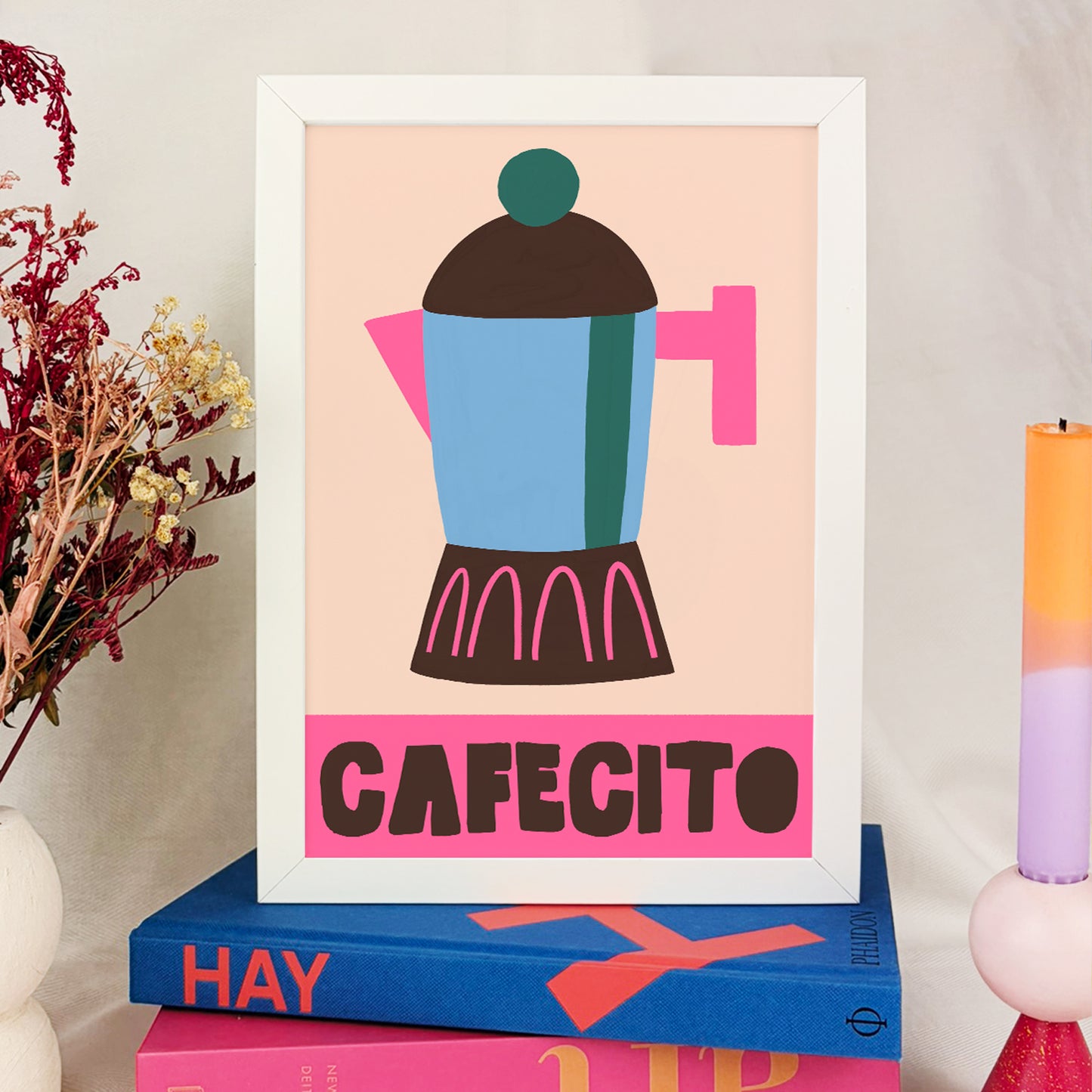 Print enmarcada en blanco de una cafetera con mango rosa y la palabra cafecito debajo con letras negras sobre fondo rosa puesta sobre una pila de catálogos de diseño