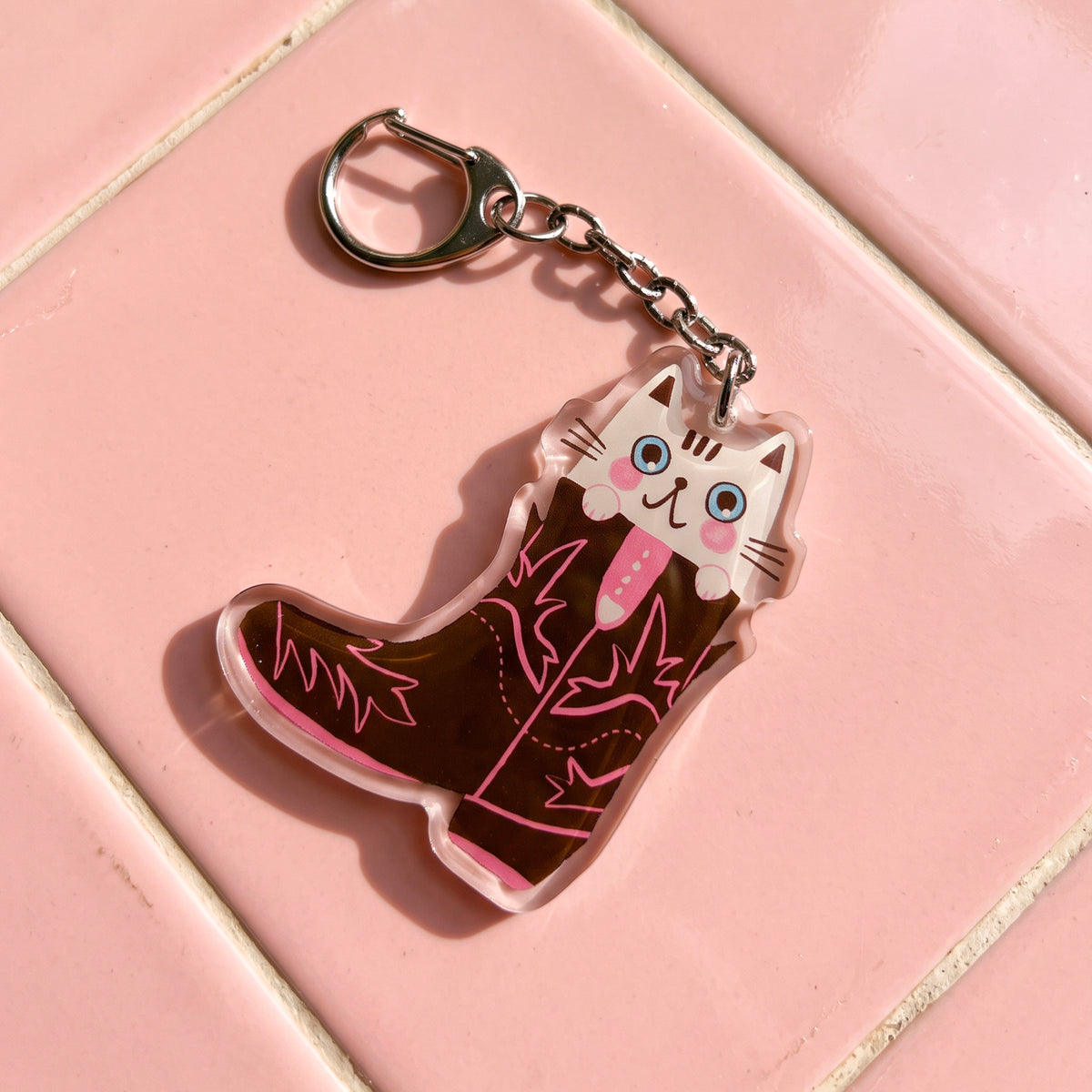 Llavero de acrílico de Laura Ortiz con forma de bota vaquera marrón y detalles rosas, con un gato blanco ilustrado asomando, sobre baldosas rosas