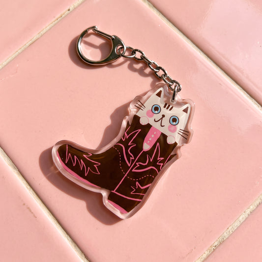 Llavero de acrílico de Laura Ortiz con forma de bota vaquera marrón y detalles rosas, con un gato blanco ilustrado asomando, sobre baldosas rosas