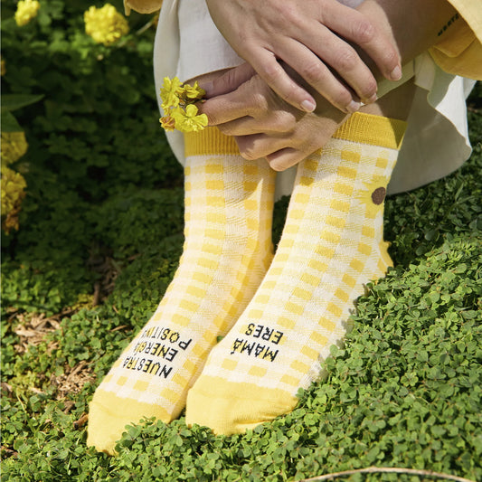 Pies sobre la hierba con los calcetines de caña baja estampados a cuadros amarillos con el mensaje "Mamá eres nuestra energía positiva"