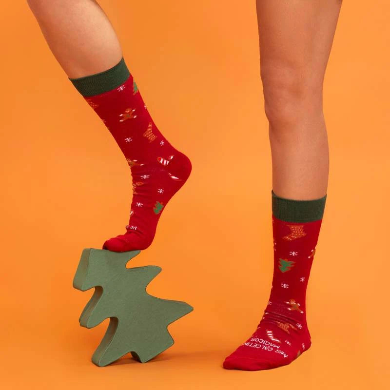 Piernas con los calcetines de navidad rojos y verdes con estampado de calcetines, galletas de jengibre, árboles, copos y bastones de caramelo pisando un árbolito de madera