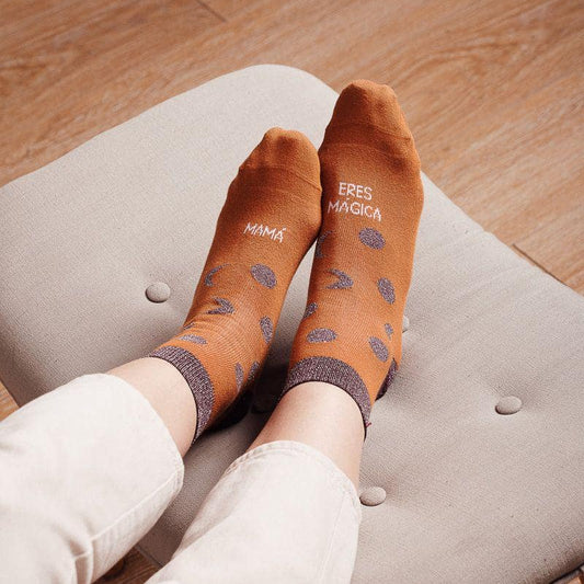 Pies en un sofá con los calcetines de caña baja marrones con detalles en purpurina y el mensaje "Mamá eres mágica"