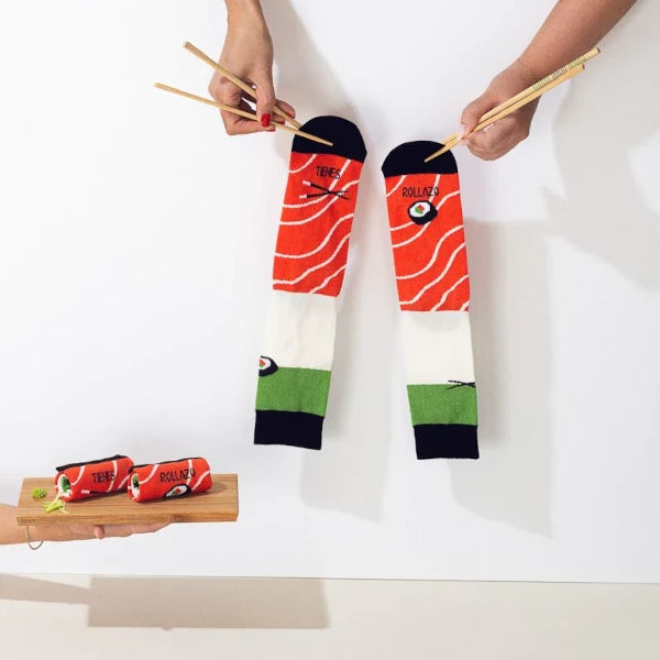 Tabla con los calcetines Tienes Rollazo de UO con los colores del sushi enrollados y dos manos cogiéndolos con palillos chinos